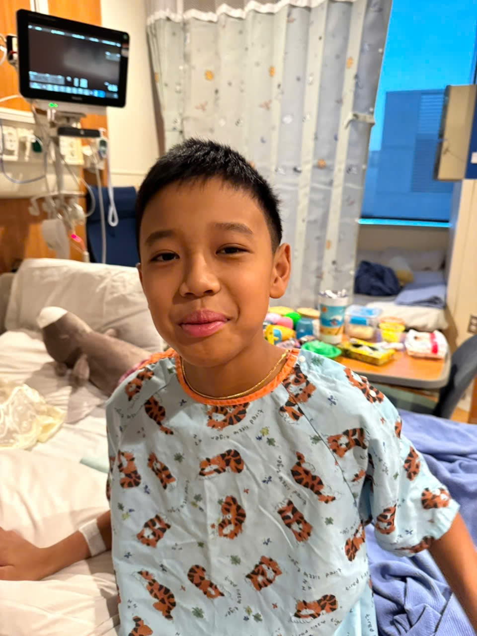 Keegan sitting up in his hospital bed in a tiger-print gown, smiling at the camera.