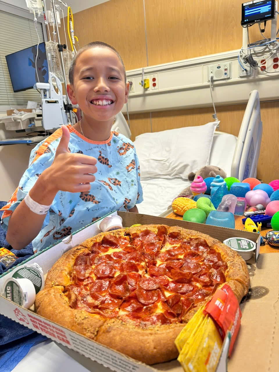 Keegan giving a thumbs up over a large pepperoni pizza on his hospital tray.