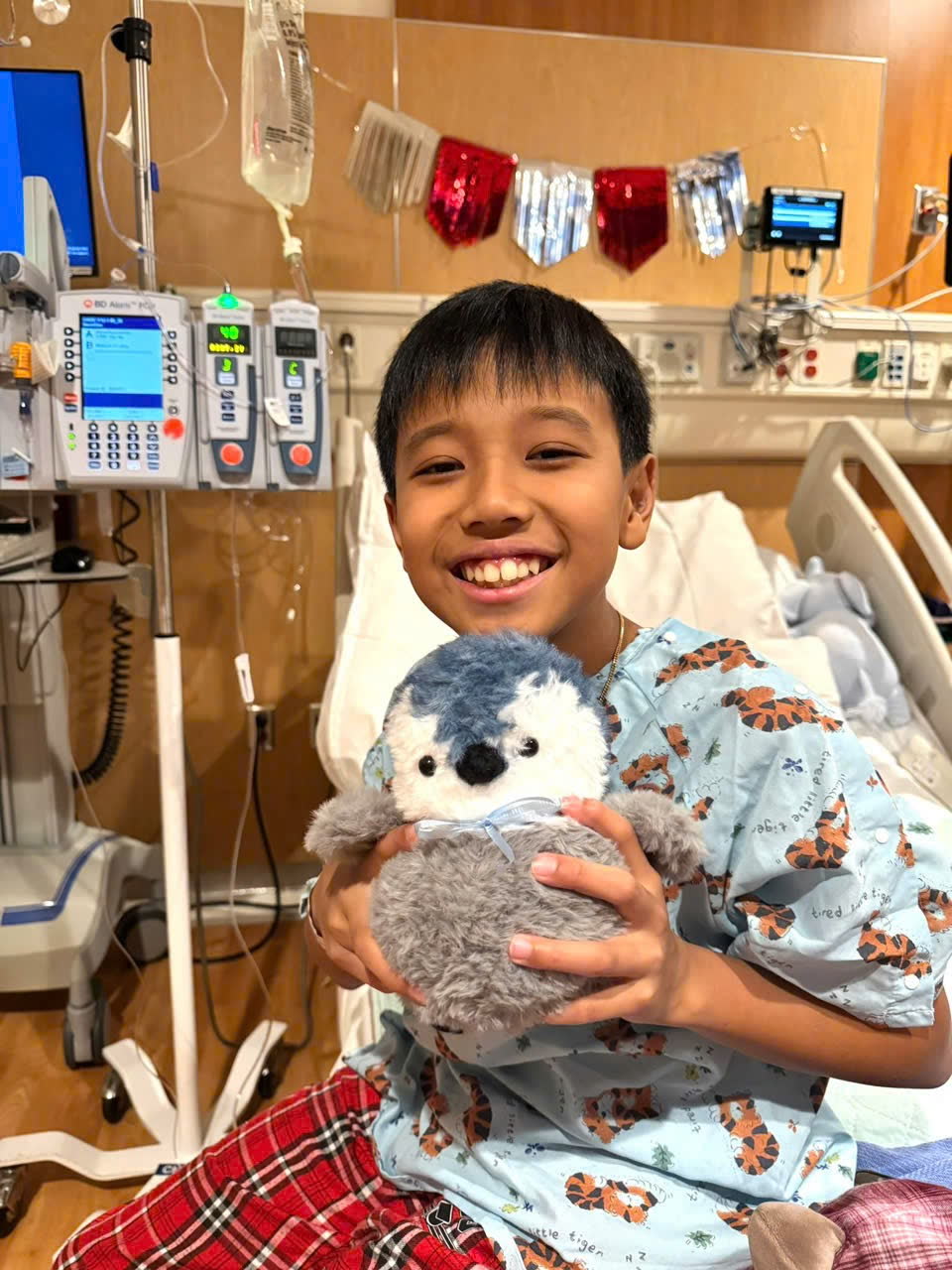 Keegan grinning while holding a stuffed penguin, with infusion pumps running behind him.